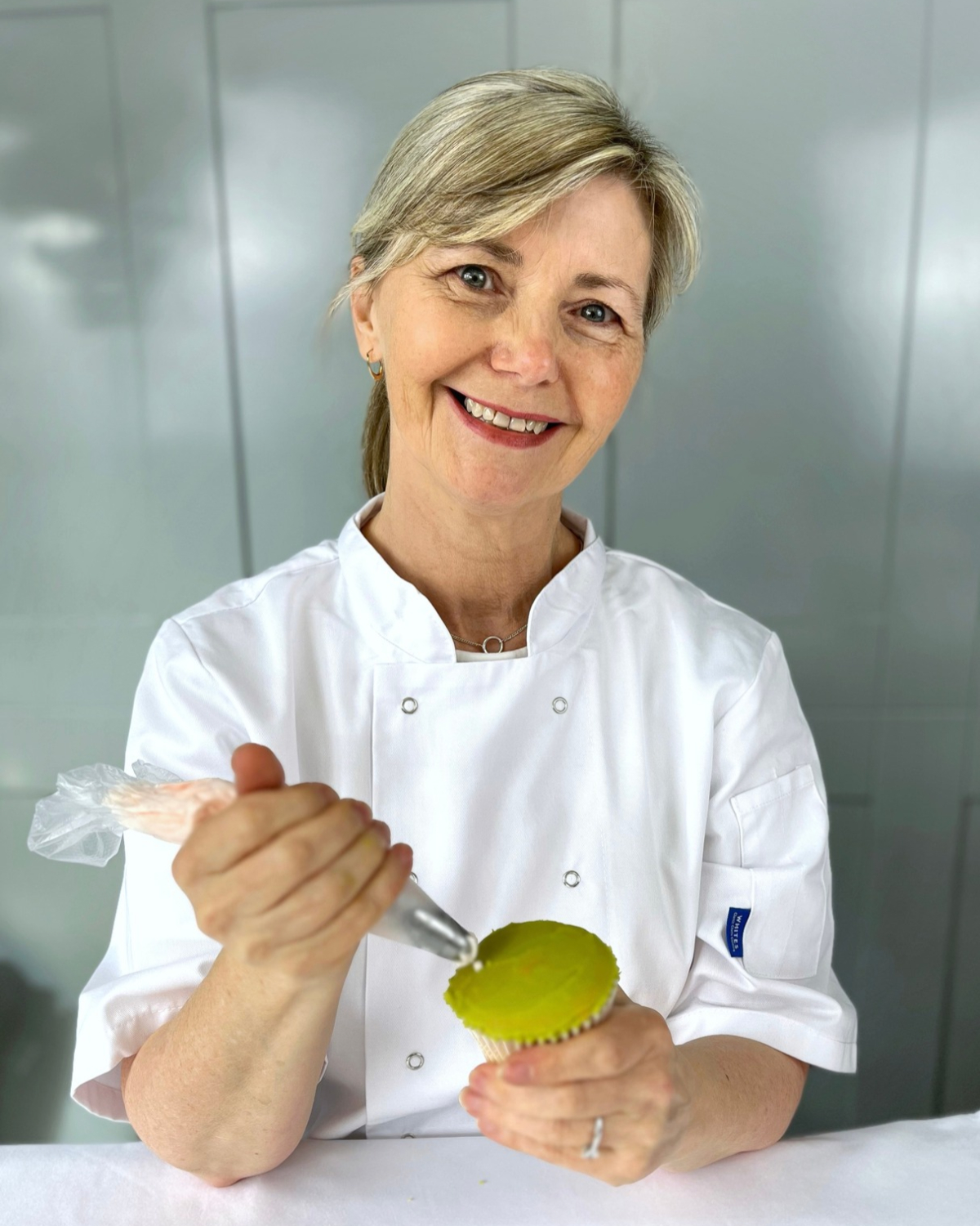 Sharon Burchall, floral cupcake artist, smiling in a white chef jacket while piping a cupcake